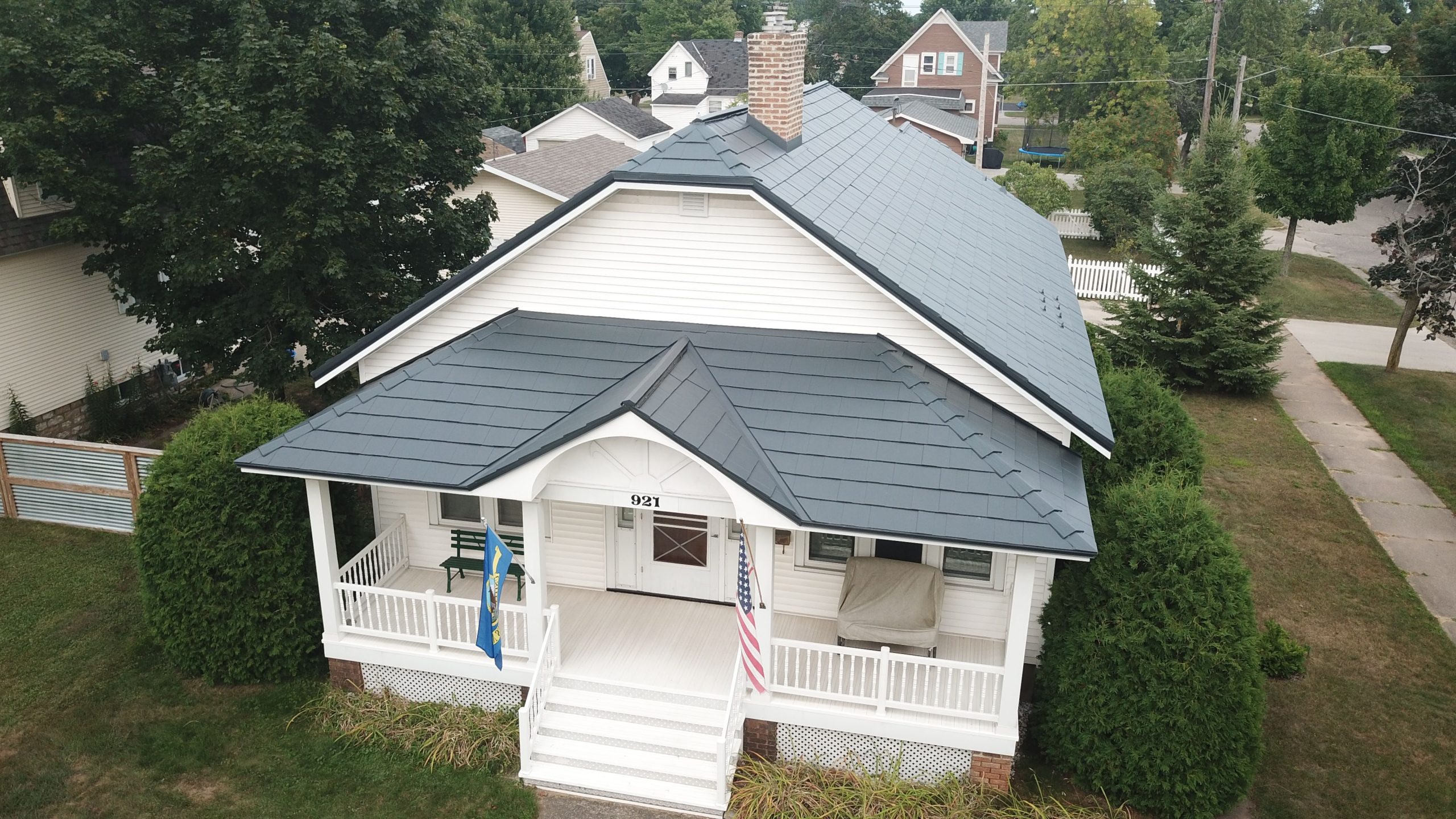 Aerial view of a classic white bungalow with a charcoal gray metal roof, a full-width covered front porch with white railings, an American flag, and mature evergreen shrubs, situated on a tree-lined residential street.